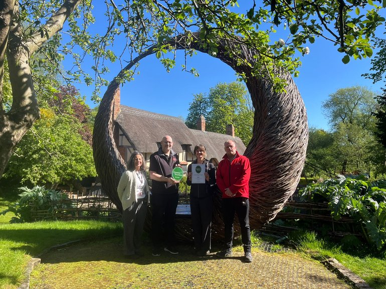 A group of SBT colleagues stood in sunshine in the garden at Anne Hathaway's Cottage in front of a willow sculpture holding the Green Tourism Award plaque and certificate.