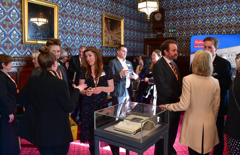 Guests gather around a display case containing historic documents and books. The room features period wallpaper, gilt-framed paintings, and ornate lighting. Attendees wear formal attire and lanyard badges, engaged in conversation around the glass exhibition case on a pink carpet