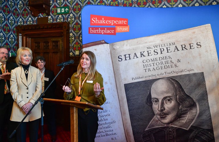 Rachael North in a mustard-coloured jacket speaks at a wooden podium with decorative period wallpaper behind her. To her right is a large display of the First Folio showing the title page and a portrait of William Shakespeare. The Shakespeare Birthplace Trust logo appears on a blue banner above.